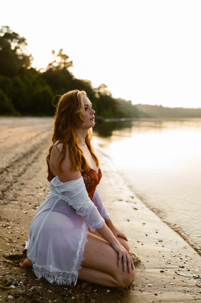 Woman in red lingerie and white robe sit on the shore for her in the water boudoir session. 