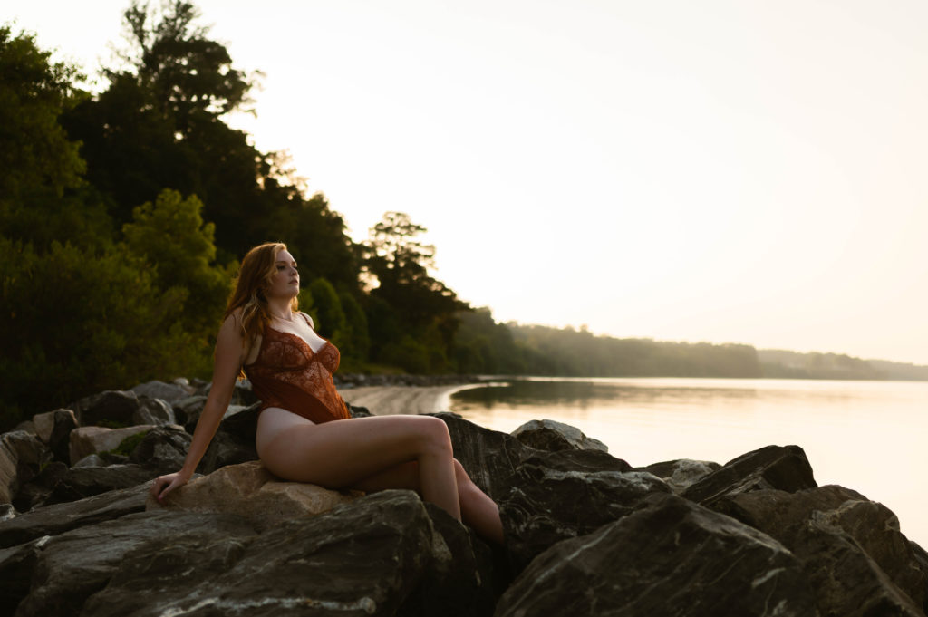 Woman in red lingerie sits on rocks next to a lake for her in the water boudoir session. 