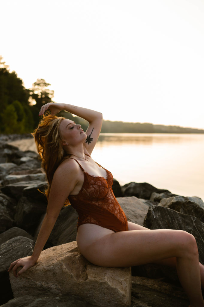 Woman in red lingerie tosses her hair back while sitting on rocks next to the lake for her outdoor boudoir session. 