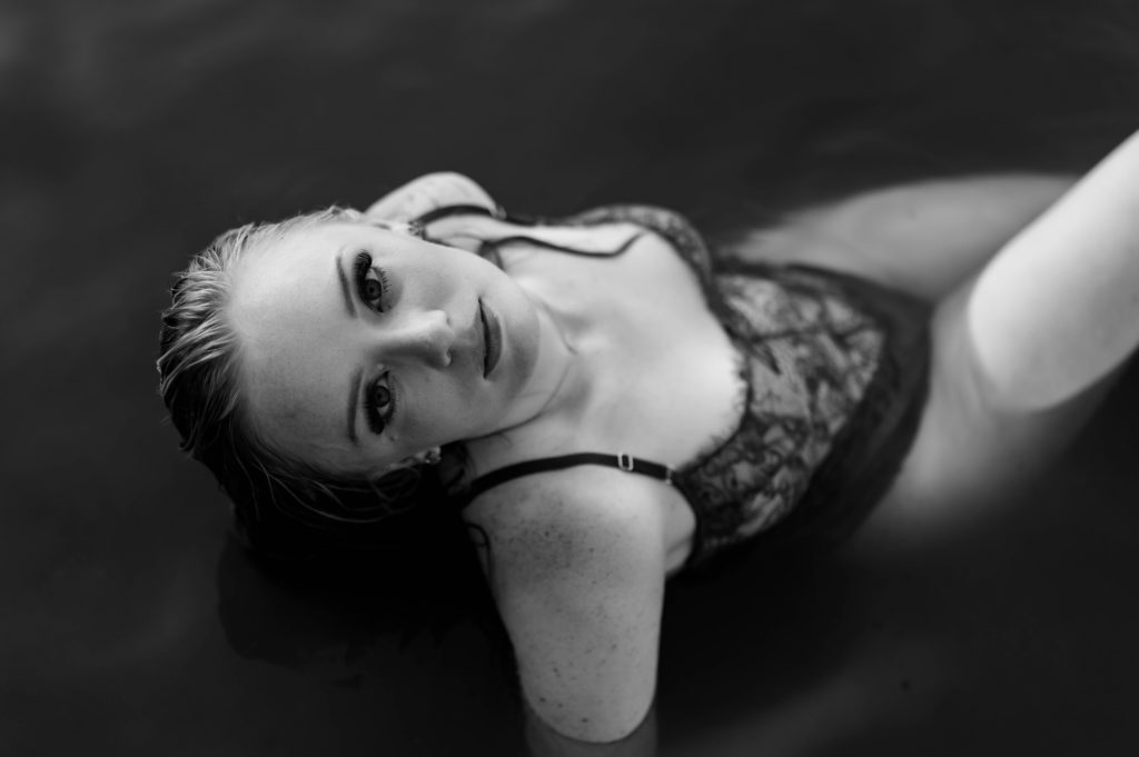 Black and white image of a woman in lingerie sitting the water looking up at the camera above. 