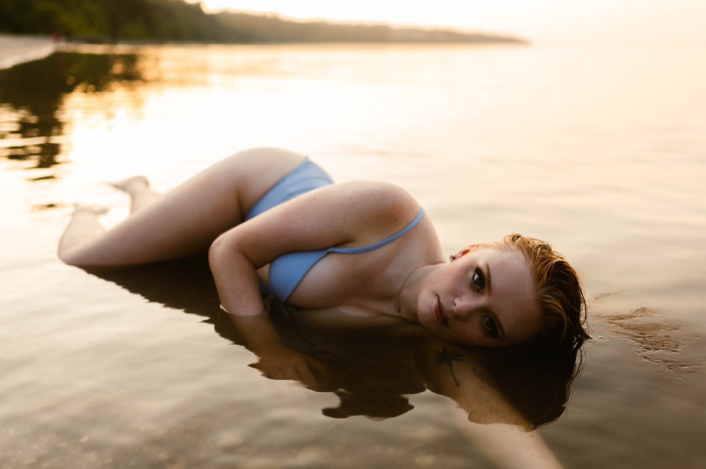 Woman in blue swim suit lies in the shallow water, looking up at the camera. 