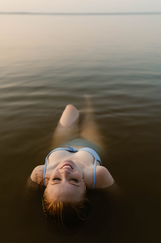 Woman in blue swim suit leans back in shallow water, smiling. 