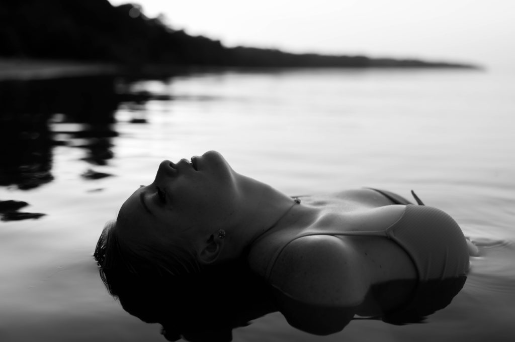 Black and white image of woman in swimsuit leaning back in the shallow water. 