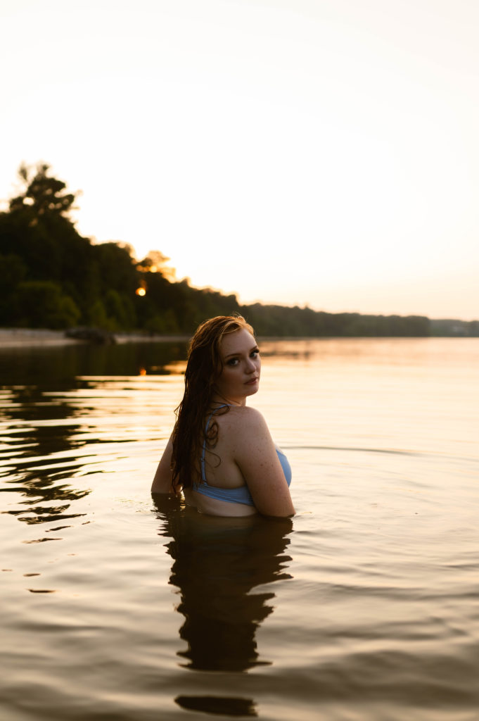 Woman in blue swim suit walking into the water away from the camera towards the setting sun. 