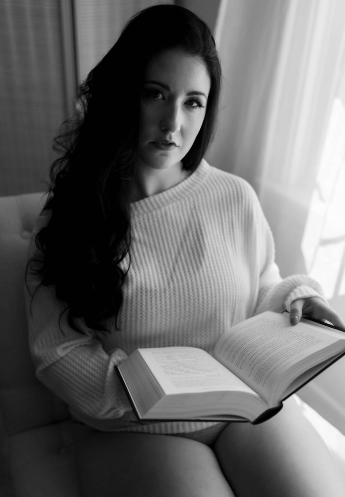 Black and white image of a woman sitting in a chair reading, looking up at the camera.