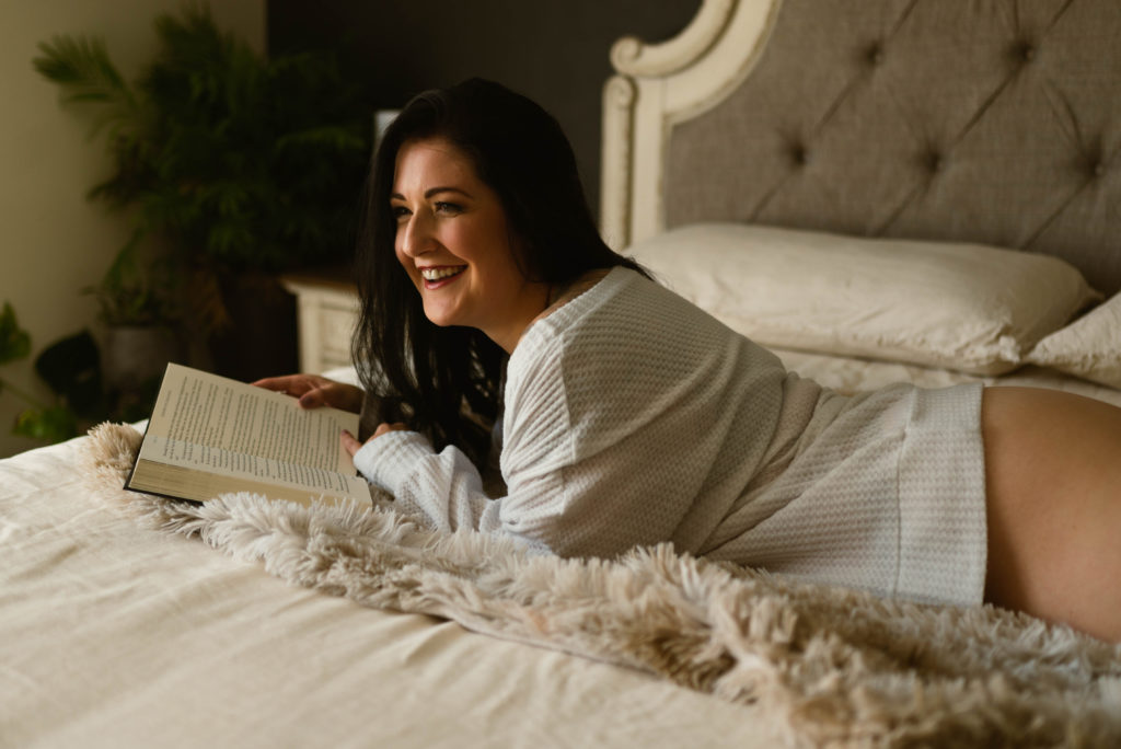 Woman in a white shirt lying across a bed reading a book at her empowering portrait session.