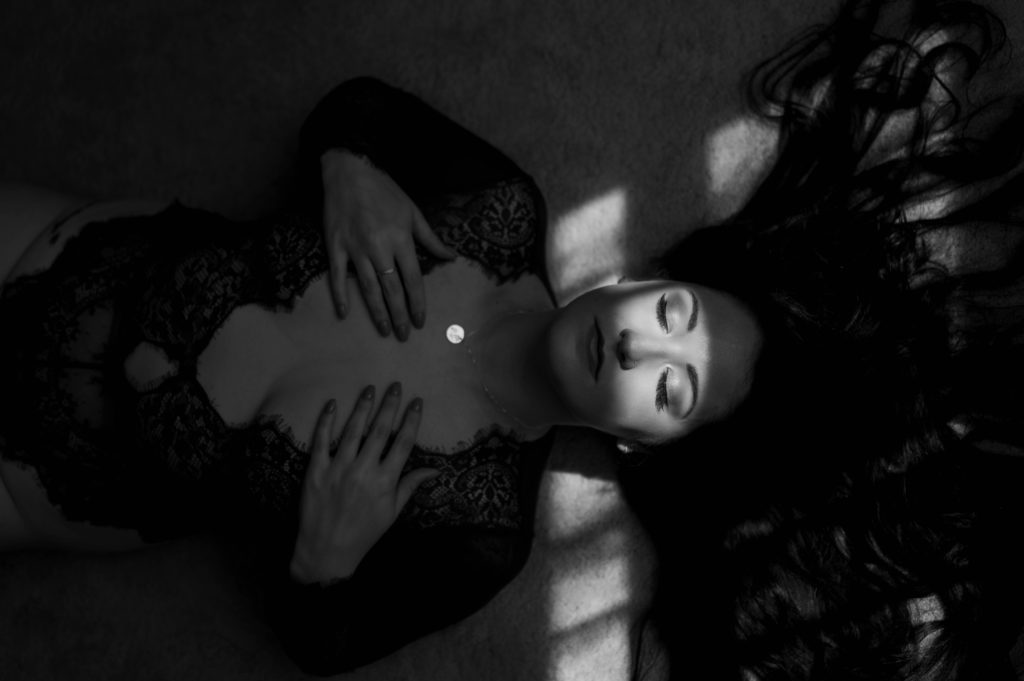 Woman in lace lingerie lying on the floor in this black and white photo with shadows from a window over her face. 