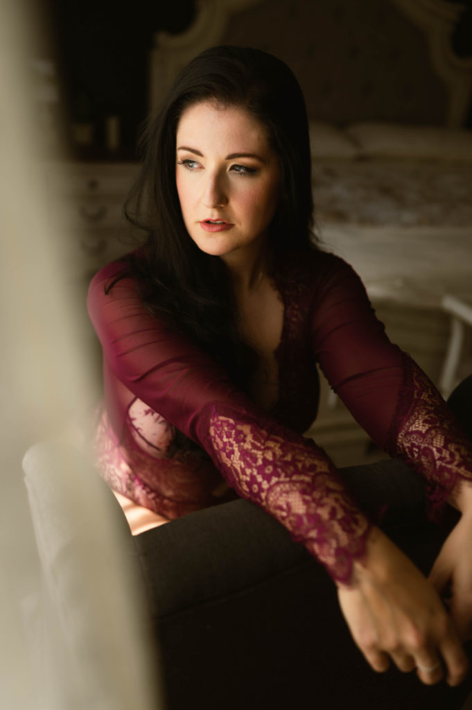 Woman in maroon lingerie kneeling over a bench looking out the window.