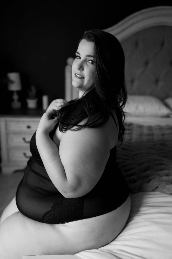 Black and white image of a woman in lingerie sitting on the edge of a bed for her boudoir session. 