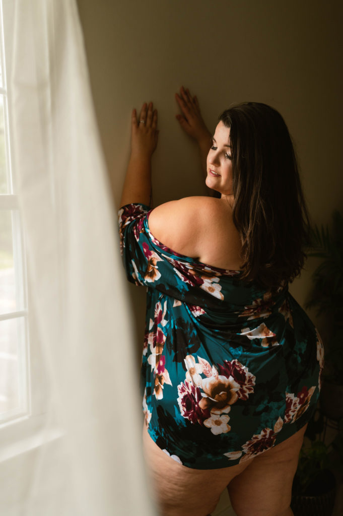 Curvy woman in a  colorful floral robe leaning against a wall and looking down to the ground. 