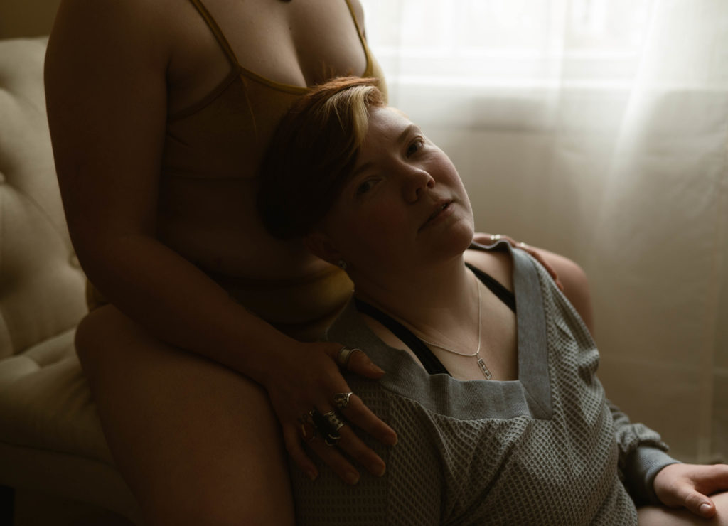 woman is leaning against her partner's legs while sitting on the floor next to a window, looking at the camera. | Virginia Sensual Portrait Photography