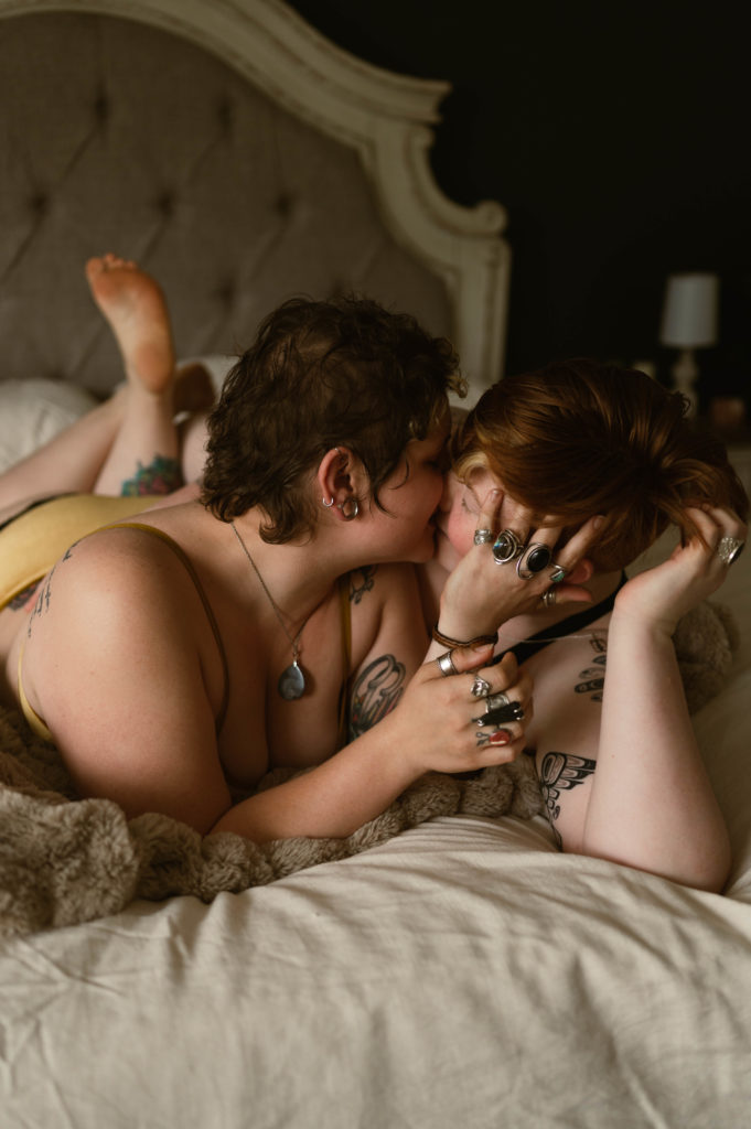 A female couple lying on the bed on their stomach, curled up close & kissing