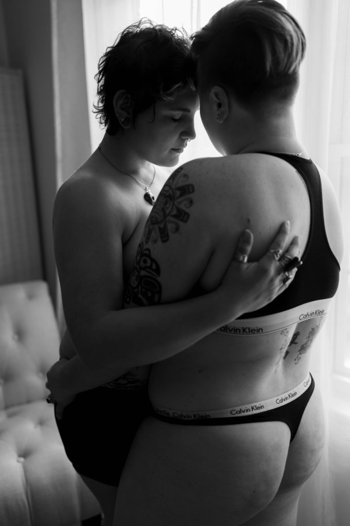 Sensual black and white photo of two women in front of a mirror in their underwear, holding each other lovingly.