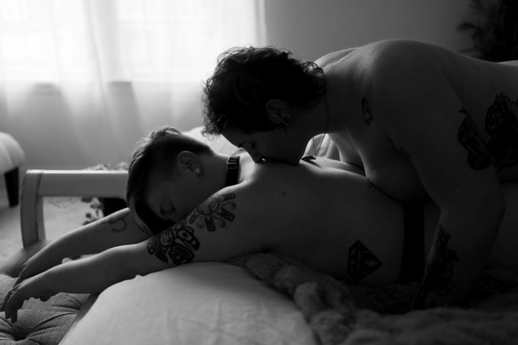 Sensual black and white photo of woman lying on her stomach on the bed while her partner lies over her back, kissing her spine.