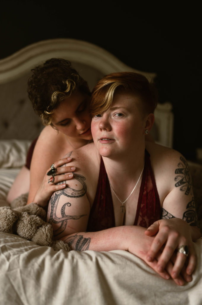 Woman with short hair is looking directly into the camera while her partner lies on her back, kissing her shoulder. | Virginia Sensual Photography