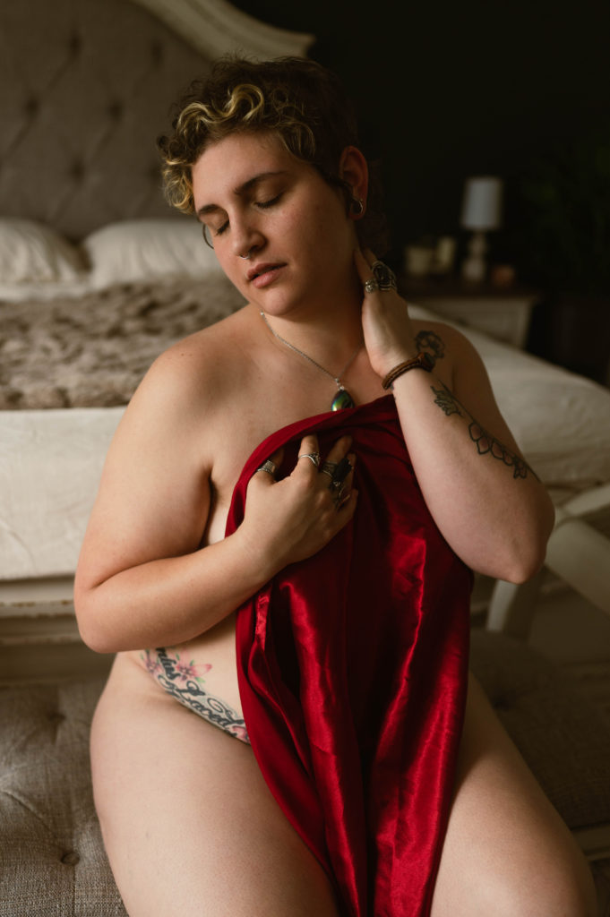 Image of a woman with many tattoos, nude and sitting at the end of a bed covering her breasts with a red silk sheet