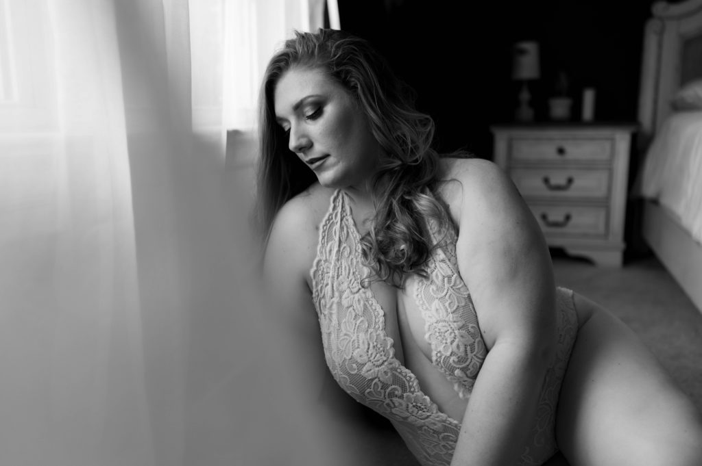 Black and white picture of a woman in lace lingerie sitting near a window. 