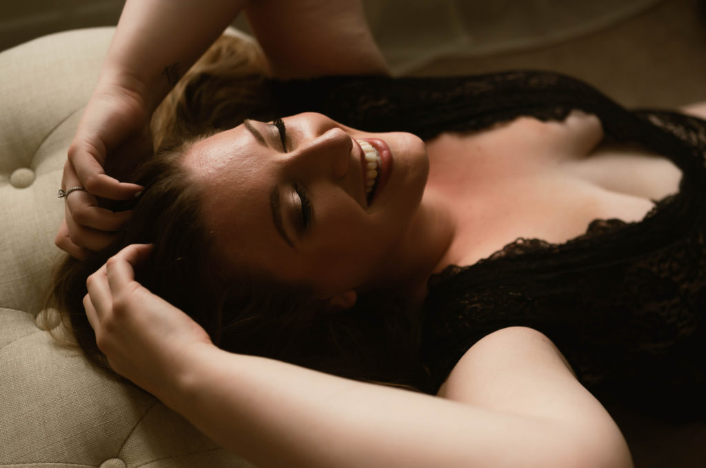 Woman wearing black lace lingerie, laughing with her hands in her hair. 