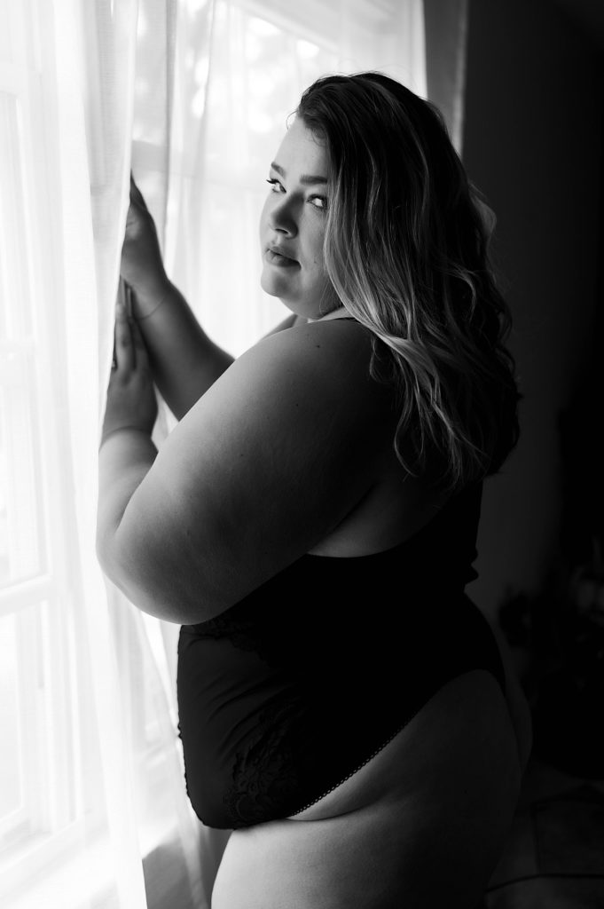 black and white boudoir photo of a plus-size woman in a black bodysuit, looking over her should at the camera.