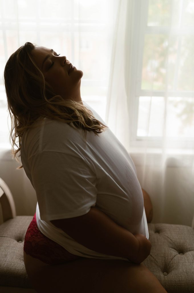 woman in a wet white tee shirt straddling a bench near a window for her boudoir session.