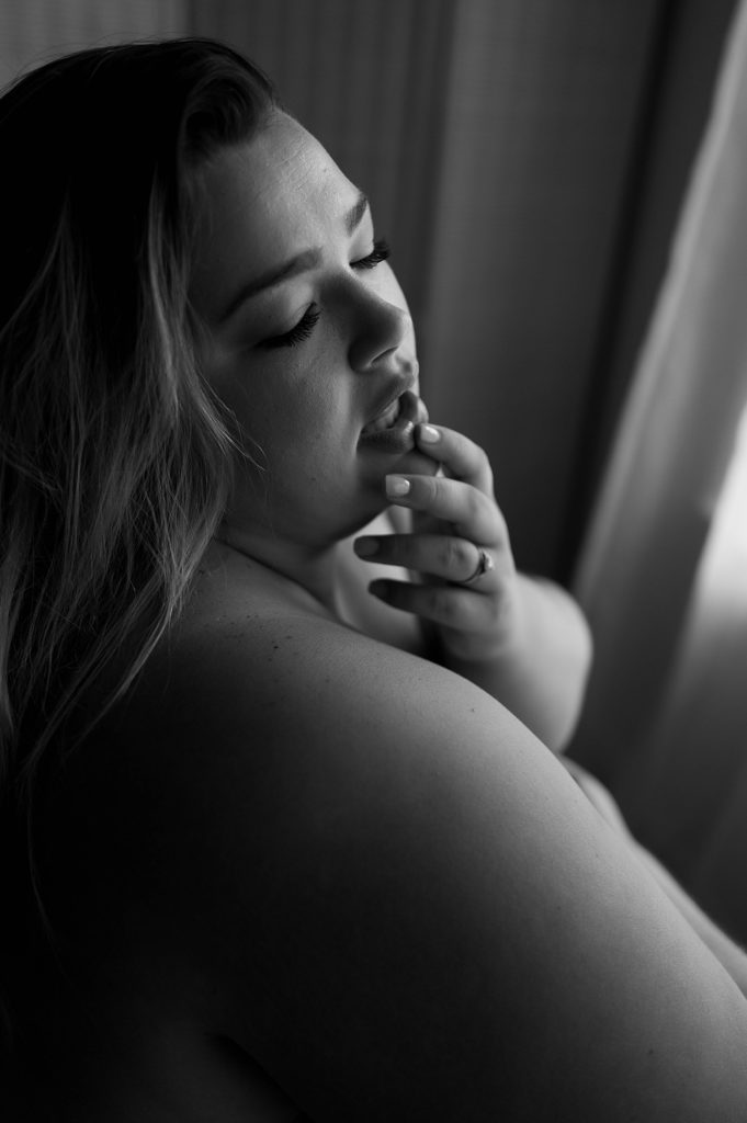 black and white boudoir image of a woman in front of a window, gently touching her lip, eyes closed. 