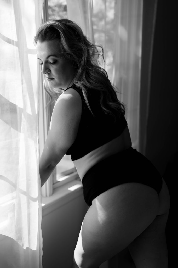 Black and white image of a woman at her boudoir session leaning against a window in black lingerie.