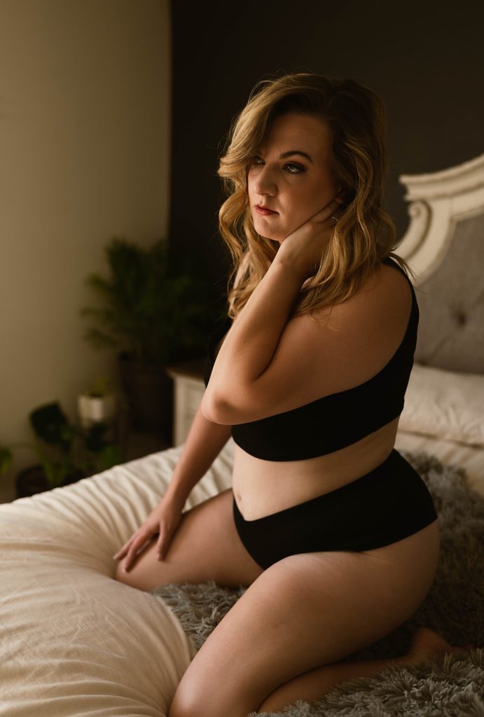 Blonde woman in a black underwear set looking into the distance while sitting on a bed at her Virginia Boudoir Session