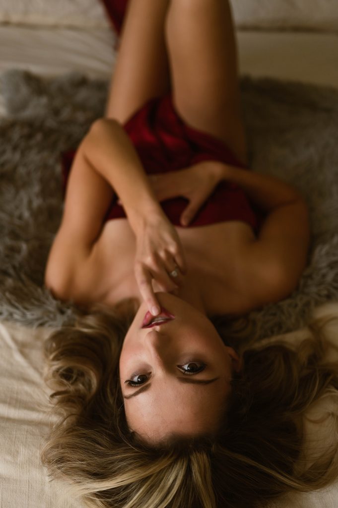 woman lying nude on a bed, covered only by a red sheet, looking back at the camera with her finger on her mouth. 