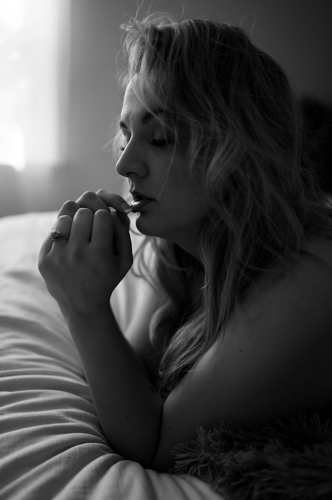 Black and white picture of a nude woman at her boudoir session, finger placed on her lip, looking down.