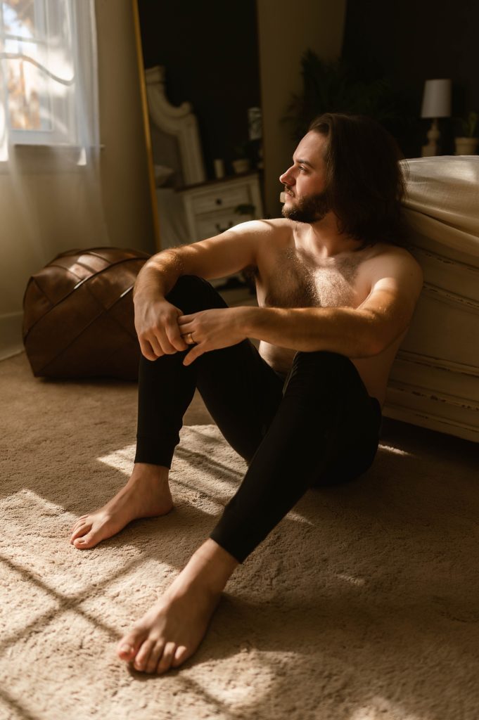 Man sitting in golden light at the end of a bed without a shirt on. 