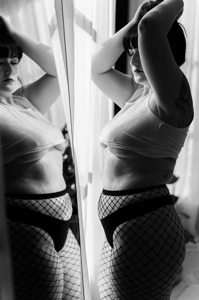 Woman in white tank top and fishnet stockings standing in front of a mirror. 