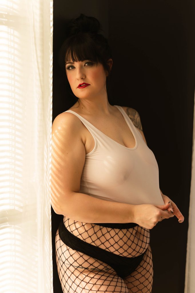 Woman in white tank and fishnets standing by a window in a boudoir studio with VA photographer, Tease by Hannah Louise