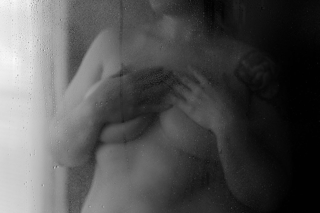 Nude woman through the glass of the shower holding her hands over her breasts. 