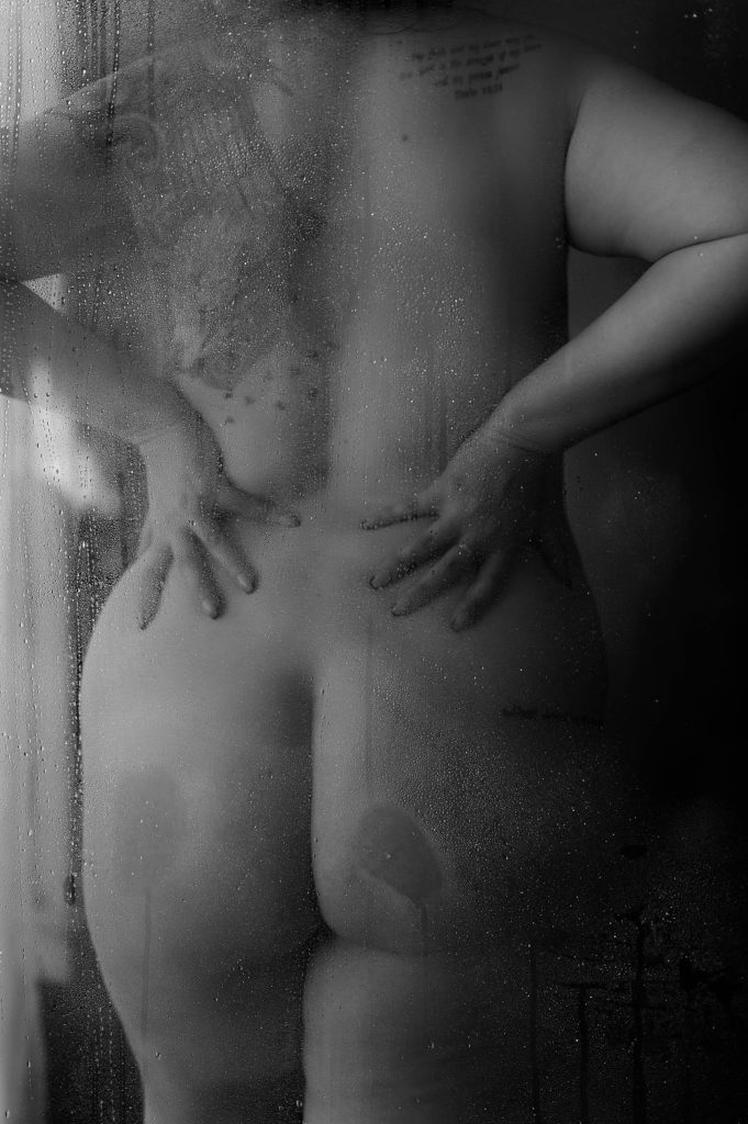 Nude woman leaning her butt against a shower glass for her boudoir session. 