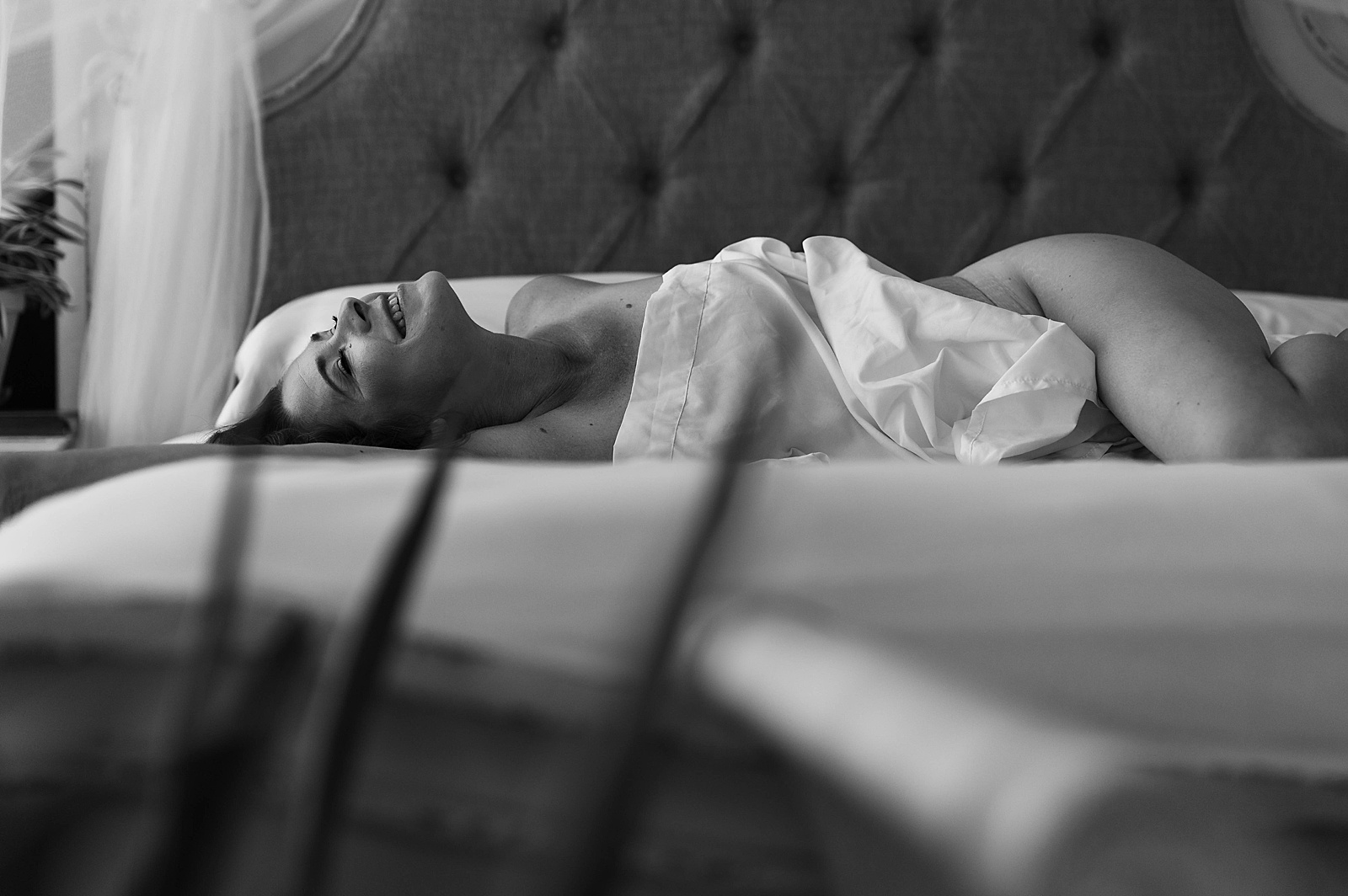 Nude woman smiling and lying across a bed covered with just the sheet 